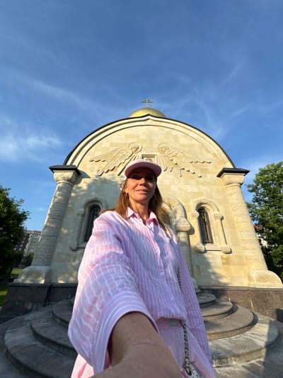 A woman stands in front of a golden-domed church in Kirishi, Russia, taking a selfie. She is wearing a pink and white striped button-down shirt and a pink baseball cap. The church is made of light-colored stone with intricate carvings and arched windows. The sky is blue with some wispy clouds, and trees are visible in the background. The woman is smiling and holding her phone in her right hand to take the picture. The perspective of the selfie is low-angle, with the church appearing large and imposing.