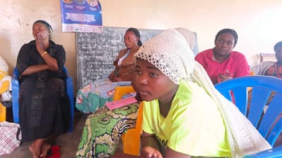 In Kamakombe, Democratic Republic of the Congo, a group of women are seated in what appears to be a classroom or community meeting space. The room has a light-colored wall and a blackboard visible behind the subjects. A poster with text and images is attached to the upper left portion of the wall.
In the foreground, a young woman wearing a lime green t-shirt and a white lace headscarf looks to her right, away from the camera. She is seated on an orange chair. Next to her, a woman in a black dress and headscarf sits on a blue chair, resting her chin on her hand. Her arms are crossed.
Behind these women, other individuals are visible. One woman wears a brown jacket over a patterned top and is looking to her left. Another woman in a pink shirt is seated and holding an object. Further back, another person is partially visible. The women are seated on various colored chairs, including blue and orange. Clothing includes patterned fabrics and head coverings. A pink folded paper is placed on the orange chair in front of the young woman. The floor is concrete. Red flip-flops are on the floor near the woman in the black dress.