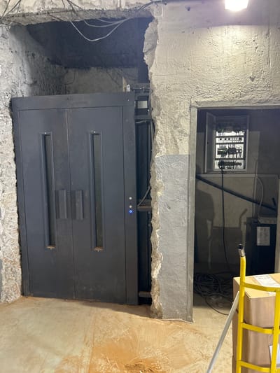 The image depicts an unfinished elevator shaft within a building under construction or renovation. A dark gray elevator door with two glass panes is prominently featured on the left. To the right of the elevator door, an open electrical panel with numerous wires and switches is visible, mounted on a concrete wall. The surrounding environment is rough and unfinished, with exposed concrete, rubble, and construction materials, suggesting an industrial or utility space. A yellow ladder and cardboard boxes are present in the foreground on the right. The scene appears to be located in Djerman, Bulgaria, as indicated by the prompt. There are no people visible in the image.