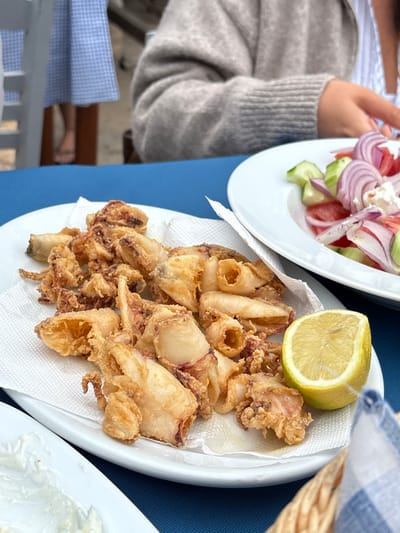 The image depicts a dining scene at a restaurant located within the Municipal Unit of Loutraki - Perachora, Greece. The foreground is dominated by a white oval plate holding a serving of golden-brown fried calamari, which rests on white paper towels. A half-cut lemon is positioned on the right edge of this plate. To the right and slightly behind, a second white oval plate contains a Greek salad, visibly composed of sliced red onions, cucumber pieces, tomato wedges, and crumbled white feta cheese. The dining table is covered with a solid royal blue tablecloth. In the bottom left, a portion of a white rectangular dish containing a creamy white dip is partially visible. A woven basket with a blue and white checkered napkin is present in the bottom right corner. Two individuals are partially visible in the blurred background. On the right, a person is seated, wearing a light gray, textured, long-sleeved garment, with their right arm and hand positioned near the Greek salad plate. Further to the left in the background, another individual is partially visible from the lower torso down, wearing a light blue and white patterned dress or skirt and dark flip-flops, suggesting they are either standing or seated further back. The overall environment suggests a casual, possibly outdoor or semi-outdoor, restaurant setting with natural light.