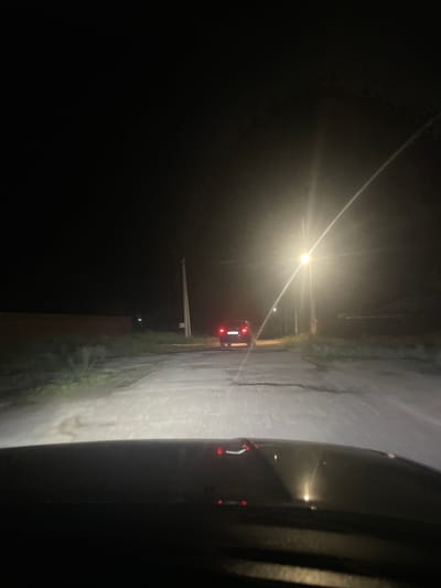 The image depicts a night scene on a road in the village of Yekaterinovka, Russia. The perspective is from inside a vehicle looking forward. A car with its red taillights illuminated is visible driving away in the distance. A bright street light illuminates the road ahead and to the right, creating a strong glare. A utility pole stands to the right of the road, near the street light. The road appears to be unpaved or in poor condition, with evidence of some patches and cracks. On the left side of the road, a low, dark structure, possibly a wall or building, is visible. The surrounding environment is dark, indicating it is nighttime, with some sparse vegetation along the roadside. The foreground shows the dashboard or hood of the vehicle from which the photo is taken, with reflections of lights visible on its surface.