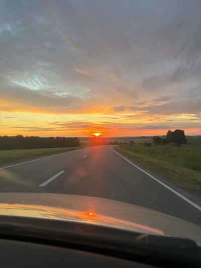 The image depicts a scenic view of a road winding through a rural landscape at sunset. The sun, a vibrant orange orb, is low on the horizon, casting a warm glow across the sky and reflecting onto the road's surface. Wispy clouds, tinged with orange and pink, stretch across the sky, creating a dramatic backdrop. The road, paved and marked with white lines, stretches into the distance, flanked by lush green fields and a dense line of trees. The foreground shows the hood of a car, with a reflection of the sunset visible on its surface. The scene appears to be located in the vicinity of Поварёнки, Russia, a region known for its picturesque countryside and natural beauty. There are no people or animals visible in the image, and the overall atmosphere is one of tranquility and natural splendor.