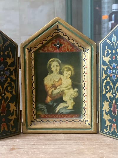 This is a close-up, eye-level, indoor shot of a triptych icon depicting the Virgin Mary holding the infant Jesus. The icon is set against a blurred background with hints of shelves and bottles. The triptych is made of wood with a distressed, golden-brown finish and intricate decorative borders. The central panel features a painting of Mary and Jesus, with Mary in a golden-orange robe and Jesus in a light yellow garment. The side panels are dark teal with gold and red decorative patterns. The icon is placed on a light-colored wooden surface, possibly a table or shelf. The location is identified as Zeneta, Spain.
