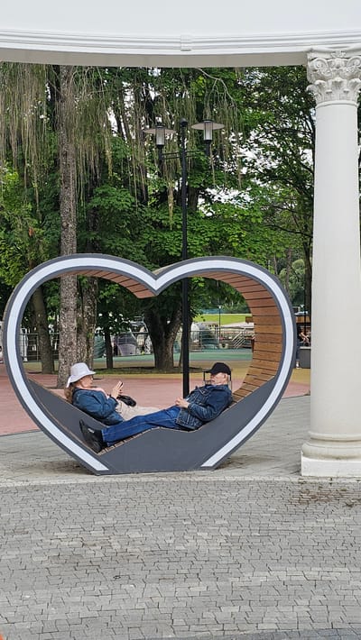 An elderly couple is relaxing on a heart-shaped bench in a park, possibly in the city of Alikonovka, Russia. The man, wearing a black baseball cap and a denim jacket, is lying down with his eyes closed. The woman, wearing a white sun hat and a denim jacket, is sitting up and looking at her phone. The bench is made of wood and metal and is situated on a paved area with trees and streetlights in the background. A large white column is visible on the right side of the image.