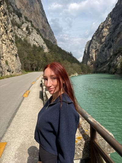An adult female subject is depicted in a medium close-up, standing on a paved surface next to a road, with the Gola del Furlo (Furlo Gorge) near Fermignano, Italy, as the backdrop. The subject, positioned slightly right of center, has long, reddish-brown hair and a fair complexion with visible freckles, smiling directly towards the viewer. She is wearing a dark navy blue, ribbed knit top with a partial silver zipper at the collar.

To her left, a paved asphalt road curves into the distance, bordered by yellow speed bumps or reflective markers along its edge. Beyond the road, a steep, rocky cliff face rises dramatically. To her right, a river with clear, turquoise-green water flows, flanked by a weathered concrete and dark metal railing barrier. Another steep, rocky cliff face ascends on the opposite side of the river. The environment is characterized by rugged limestone cliffs, sparse green vegetation on the lower slopes, and a partly cloudy sky overhead with patches of blue. Natural light illuminates the scene, casting subtle shadows from the barrier and the subject onto the ground.