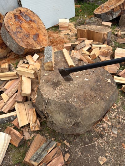 In Kapreshumi, Georgia, a scene of woodcutting is depicted. A large tree stump, measuring approximately one meter in diameter, serves as the central object. An axe with a black handle is embedded in the stump. Scattered around the stump are numerous pieces of cut firewood, varying in size and shape, suggesting recent activity. In the background, larger logs and sections of trees are visible, some of which appear to have been recently felled. A portion of a white structure, possibly a shed or building, is present in the upper left of the frame. The ground is covered with dirt, grass, and wood chips, indicative of an outdoor environment where lumber is processed. There are no people visible in the image.
