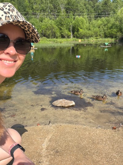 The image is a selfie taken by a person wearing a leopard-print hat and sunglasses, smiling at the camera. They are sitting by the edge of a clear, shallow body of water with a sandy bottom. Several ducks are swimming in the water in the foreground. In the background, there is a lush green forest and a calm body of water with some colorful objects floating on it. The image depicts a sunny day in a natural outdoor setting, likely a park or a recreational area. The location of Кульдур, Russia is not evident from the visual information provided in the image.