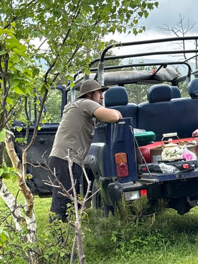 A person wearing a green t-shirt and a wide-brimmed hat leans against the back of a dark blue off-road vehicle in a grassy, wooded area. The vehicle's rear is open, revealing various items including a white teapot with green floral designs, a green cushion, and other bags. The background shows trees and a cloudy sky, suggesting a natural, possibly mountainous, environment. The image location is identified as Nizhnyaya Teberda, Russia.