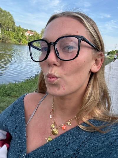 A woman is shown in a close-up, outdoor shot. She is wearing black glasses, a blue sweater, and a gold necklace with charms. She has blonde hair and is making a kissing face. In the background, there is water, grass, trees, and a building. In the distance, two people are walking on a path. The photo was likely taken in Marquette-lez-Lille, France, given the presence of typical architecture of the region.
