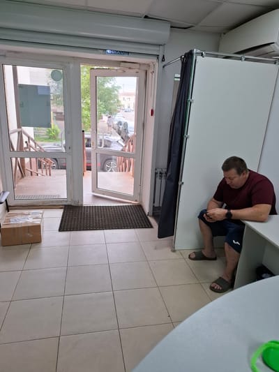 A man is hunched over in a maroon t-shirt and dark blue shorts, his face obscured as he appears to be working or looking at something on the floor. He is wearing sandals. The scene is indoors, with tiled flooring and white walls. A closed door with glass panels is visible, and through the glass, an outdoor scene with trees and buildings can be seen, suggesting it is daytime. There are two cardboard boxes stacked near the door. A dark doormat is placed in front of the door. The overall impression is of a simple, everyday setting.