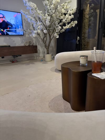 This image depicts a modern living area within a residence located in Mougins, France. The perspective is from a low angle, looking across a light-colored floor towards a wall-mounted television and various furnishings. On the left, a large flat-screen television displays a close-up of an individual, likely female, wearing sunglasses and a headset in what appears to be a vehicle's cockpit. Below the television, a floating dark wooden console holds two light-colored, possibly ceramic, circular decorative objects and a stack of books, including one titled "ROLEX." To the right of the console, a tall artificial cherry blossom tree with dense white flowers stands in a white cylindrical planter. The floor consists of a light grey polished surface, with a rectangular segment covered by a light-beige textured rug in the foreground. Beneath the console, a small red, white, and blue toy car and a small blue ball are visible on the polished floor. In the mid-ground right, a white, rounded sofa is partially visible. In the immediate foreground, two dark bronze-toned, organically shaped tables are present. The larger table displays a white Jo Malone brand candle in a clear glass container with a silver lid. The smaller table holds a clear plastic cup with a straw containing a reddish-brown liquid, alongside a white napkin. Beyond the artificial tree and sofa, a large glass panel reveals an exterior view of a textured stone wall.