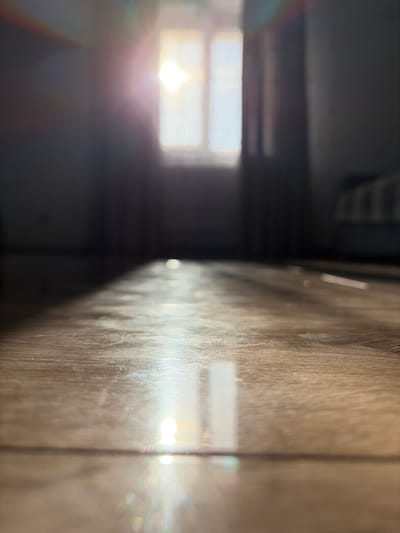 The image depicts a low-angle view of a room with natural light streaming through a window. The floor appears to be made of wood or a similar material with a visible grain pattern. Sunlight creates a bright, sharp reflection across the floor, with subtle prismatic effects indicating the presence of dust particles in the air or imperfections on the floor's surface. In the background, a window with blinds or curtains is visible, with the bright light of the sun partially obscuring the details outside. The overall impression is one of a quiet, interior space illuminated by morning or afternoon sun. There are no people or specific objects that suggest a particular location within the city of Hanniv, Ukraine, other than the generic elements of a residential or office interior.