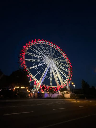 The image depicts a large Ferris wheel illuminated with red lights against a dark night sky. The Ferris wheel is centrally located and appears to be in operation, with some gondolas visible. In the foreground, a road is visible with lane markings. To the left of the Ferris wheel, there are trees and some illuminated structures, suggesting an amusement park or fairground setting. The scene is lit by the lights of the Ferris wheel and some streetlights, casting a dark, atmospheric glow. The Ferris wheel is situated in Neuss, Germany, at the "Kirmesplatz" on the Kaiser-Friedrich-Ring.