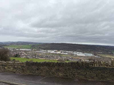 The image presents a panoramic view across a valley within the city of Calderdale, United Kingdom, captured under heavy overcast conditions. In the immediate foreground, the edge of a dark asphalt road surface is visible at the bottom of the frame, bordered by a dry-stone wall composed of irregular grey and brown masonry. Beyond this wall, the midground encompasses a developed landscape featuring green fields interspersed with residential areas comprising numerous closely-packed terraced and semi-detached houses, identifiable by their dark roofs. Adjacent to the housing, a commercial or industrial zone is present, characterized by several large, low-rise structures with lighter-colored roofs. A discernible thoroughfare runs through the valley floor. The background is dominated by undulating hills, densely covered with what appears to be deciduous woodland, currently devoid of foliage. Several tall, slender structures, consistent with communication masts or power pylons, are visible atop these distant hills. The sky is entirely covered by a uniform layer of thick, grey clouds. No individuals, animals, vehicles, specific actions, or interactions are observable within the depicted scene.