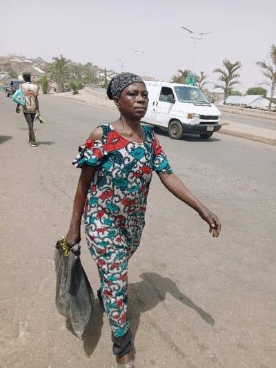 The image portrays an outdoor daytime scene on a paved road in Kadoma, Zimbabwe. A dark-skinned woman occupies the foreground, walking towards the viewer's left with her gaze directed slightly left of the camera. She wears a patterned off-the-shoulder jumpsuit or dress, featuring red, teal, and white floral or abstract designs, and a dark, possibly patterned headwrap. Her left hand clutches a dark-colored bag containing a bright yellow item. Her right arm is extended forward, slightly bent.

In the midground, a male figure walks further down the road, away from the camera, carrying light blue/green plastic items in his left hand and a light-colored object in his right. A white, high-roof commercial van, resembling a Volkswagen Transporter, is visible on the opposite side of the road, facing away from the camera. Several solar-powered streetlights are erected along the thoroughfare.

The background consists of dusty, sparsely vegetated hills dotted with residential structures. The sky overhead appears hazy and light, and direct sunlight casts distinct shadows of the subjects and objects on the asphalt road surface.