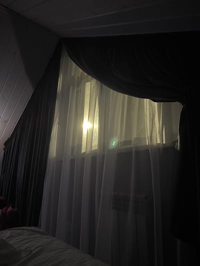 The image depicts an interior scene, likely a bedroom, illuminated by a soft, warm light emanating from a window covered by sheer white curtains. The curtains are drawn, and the light appears to be from an external source, possibly the sun or artificial lighting, creating a diffused glow. Behind the sheer curtains, a darker, heavier curtain is visible, suggesting a layered window treatment. The ceiling appears to be sloped and paneled with wood, common in attic or dormer rooms. A radiator is faintly visible behind the sheer curtains. In the foreground, the corner of a bed with striped bedding is visible, indicating the vantage point of the photograph. The overall ambiance is serene and dimly lit, with a focus on the interplay of light and fabric. No people or specific actions are depicted. The location information provided, "Doma 1096 Km, Russia," is too vague to provide specific environmental context beyond a general indication of a residential setting in Russia.