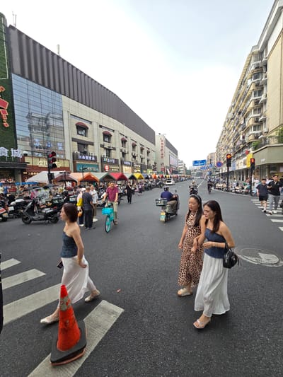 In the city of Gongshu District, China, on a cloudy day, a busy street scene unfolds with pedestrians and vehicles. The street is lined with multi-story buildings, some with glass facades reflecting the sky and surrounding architecture, and others with numerous windows and air conditioning units. A traffic light with red and yellow signals is visible, indicating a controlled intersection. Several people are walking on the pavement and crossing the street. Two women in long white skirts and dark tops are walking together, one holding the arm of the other. Another woman in a leopard-print dress is also walking in the foreground. A man is riding a blue bicycle with a basket in front. Numerous motorcycles and scooters are parked on the side of the road and being ridden by people. Several makeshift stalls with awnings are set up along the sidewalk, suggesting a market or food vendors. The road itself is asphalt, with white pedestrian crossing lines. An orange traffic cone is placed on the pedestrian crossing. The overall impression is one of urban commercial activity and pedestrian traffic.
