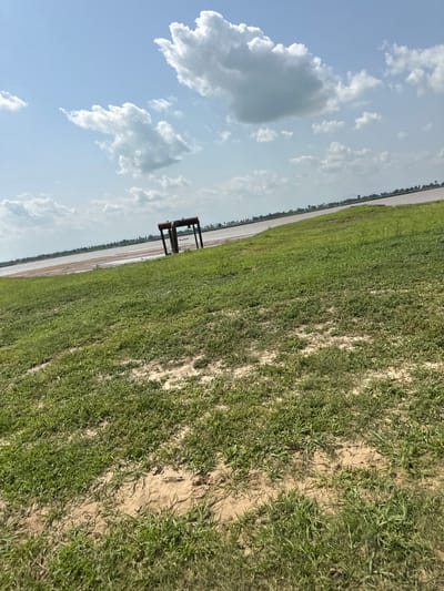 The image captures an outdoor scene in Lau, Nigeria, likely during the daytime with a bright, cloudy sky. The perspective is tilted, giving a dynamic feel to the landscape. The foreground is dominated by a grassy field with patches of exposed soil, suggesting dry or sandy terrain interspersed with vegetation.

Further into the image, a body of water is visible, appearing somewhat muddy or murky, bordering the grassy area. A row of trees can be seen along the distant edge of the water. In the middle ground, a dark, rectangular metal structure, possibly a bench or table, stands prominently on the grass. Its simple design and dark color contrast with the natural surroundings.

The sky above is a light blue, filled with scattered white cumulus clouds. The lighting suggests it is a clear day, though the clouds indicate potential for varying weather. The overall impression is one of a rural or natural setting, perhaps a park, recreation area, or the edge of a natural landscape near a body of water. No people are visible, and no text is present in the image. The scene evokes a sense of tranquility and open space.
