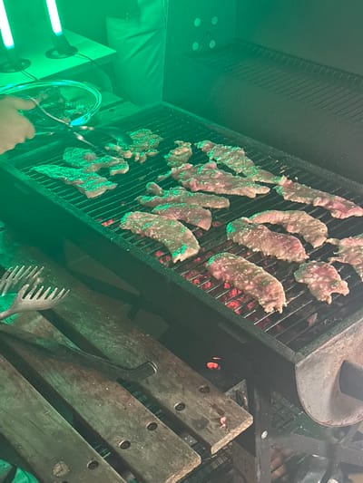 Meat cooks on a grill with hot coals underneath. Tongs and a fork are visible next to the grill. The meat is light pink, glistening with moisture, and appears to be in narrow strips. The grill itself is made of black metal. Some wooden boards are located next to the grill. The lighting in the scene is primarily green.