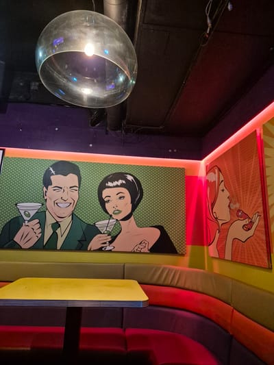 The image shows a portion of a brightly lit interior space, likely a bar or lounge, with a focus on colorful artwork and seating.
On the right is a section of curved seating, featuring alternating colors of yellow, beige, red, and purple/burgundy upholstery. On the walls are two framed, comic-book-style art pieces. The one above depicts a person smoking a cigarette or similar item, while the one below shows a man and a woman holding martini glasses against a green polka-dot background.
To the left, there is a large, spherical light fixture with a bulb inside. The background is predominantly dark, with hints of black and purple walls.
While specific details connecting it directly to Cardiff, United Kingdom are not discernible from the image alone, it appears to be a trendy or stylized venue with an emphasis on vibrant decor.