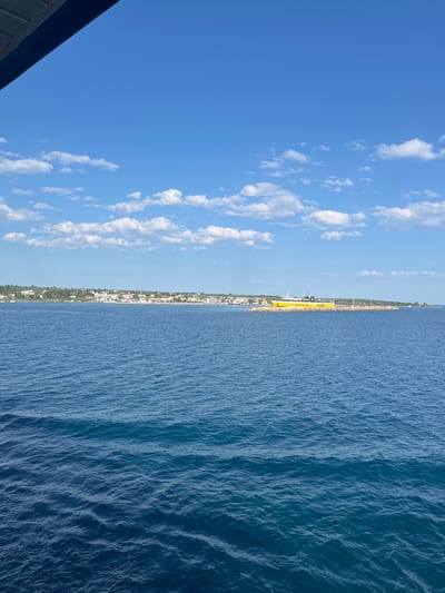 This is a wide-angle, eye-level shot taken from a body of water looking towards a coastline under a bright blue sky with scattered white clouds. In the foreground, the deep blue water exhibits small waves. The midground shows a distant coastline with a town or village featuring white buildings nestled amongst green foliage. A prominent feature in the midground is a long, yellow ferryboat docked at a breakwater. The ferry has the text "Levante Ferries" and the number "11880" visible on its side. The far background consists of the horizon line where the sea meets the sky. A dark, angular structure, possibly the edge of a boat or roof, is visible in the upper left corner of the frame. The scene appears to be a view of the Municipal Unit of Kastro - Kyllini, Greece, likely from the sea.