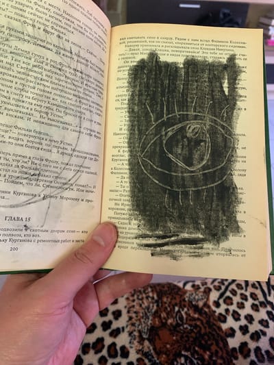 In this image, a person's hand is holding open a book, revealing text and a charcoal drawing on the right page. The drawing appears to be an abstract design with circular elements and perhaps a stylized face within them. The text on both pages is in Russian. The book itself appears aged and well-read. The background shows a person's leg covered by a garment with a leopard print. The setting suggests an indoor environment, possibly a home, with the book being the central focus of attention. The lighting is soft, indicating it could be daytime or artificial lighting. The overall mood is quiet and contemplative, centered around the act of reading and interacting with the book's content. The visible text includes chapter titles and narrative passages, providing context for the literary work.