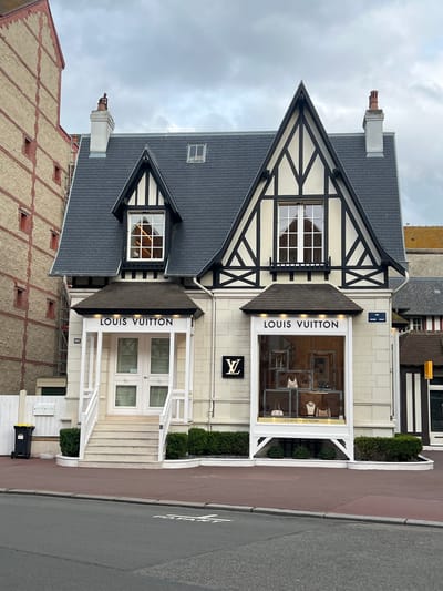 The exterior of a Louis Vuitton store in Deauville, France, is depicted. The building has a distinctive Tudor-style facade with black timber framing against a cream-colored wall. It features a prominent gabled roof and a large display window showcasing handbags and jewelry. The store entrance is marked by a white portico with stairs leading up to double doors, and illuminated "LOUIS VUITTON" signage is visible above the entrance and on the display window. Adjacent to the store, a beige building with red brick accents is visible. The street in front of the store is paved with asphalt and bordered by a red sidewalk. A black trash bin with a yellow lid is situated to the left of the store entrance. The sky is overcast.