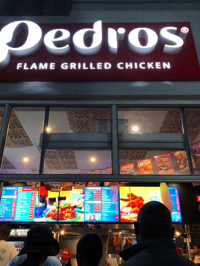 A street view shows the signage for “Pedros FLAME GRILLED CHICKEN” illuminated vertically in white against a red background. Adjacent is a window displaying menu boards with bright images and price lists, including specials like “CAR Minil” and "NEW Chilli." Some people are standing outside the restaurant, possibly looking at the menu boards. The restaurant's interior design is visible through the window, featuring a decorative pattern.