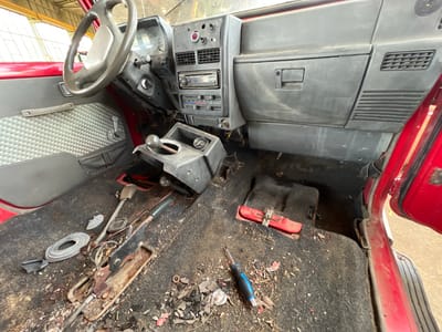 The image shows the interior of a red vehicle, likely a Suzuki Samurai or similar off-road utility vehicle, in a state of disarray. The floor is covered in a dark, worn carpet with a significant amount of debris, including leaves, dirt, small pieces of plastic and metal, and what appears to be a discarded screwdriver. The dashboard is visible, featuring a radio, various switches and knobs, and air vents. A steering wheel is present on the left side of the image. A gear shifter is mounted on the center console. The handbrake lever is also visible on the floor. The door panel on the left has a distinctive patterned insert and a manual window crank. The overall impression is one of neglect and disuse. The image was taken indoors, possibly in a garage or workshop, suggested by the lighting and background structures. The provided metadata indicates the location as Lanžhot, Czechia. There are no people visible in the image, and no actions or interactions are depicted.