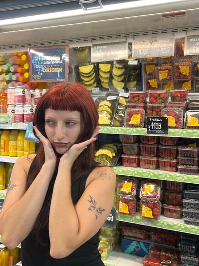 A young adult female is depicted standing in the produce section of a supermarket located in Quilmes Oeste, Argentina. She has long, reddish-brown hair with straight bangs, light eyebrows, a septum piercing, and dark lipstick, wearing a black sleeveless top. Visible tattoos include a stylized insect or butterfly design on her left upper arm and multiple small star shapes on her left shoulder. She is striking a pose with both hands raised to either side of her face, fingers slightly spread, with her gaze directed slightly to her left. The background consists of brightly lit refrigerated display shelves. To the left, various bottled and canned beverages are visible, along with a sign reading "cuidadas DESDE SU ORIGEN" with a banana icon. To the right, multiple tiers of packaged fresh produce are displayed, predominantly clear plastic containers of strawberries labeled "FRUTILLA" and priced at "$ 1.199", alongside pre-cut pineapple slices in clear plastic trays priced at "$ 4.299". Other berries and general snack fruit containers with visible pricing labels are also present on the shelves, indicating a commercial grocery environment.