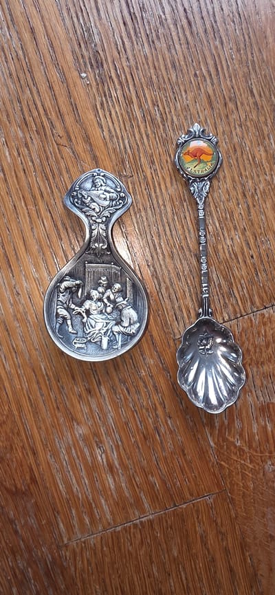 Two decorative spoons are displayed on a wooden surface. The spoon on the left has an ornate handle with an embossed scene of people gathered around a table, with one person playing a lute at the top. The spoon on the right has a small circular embellishment at the top depicting a kangaroo with the word "AUSTRALIA" below it. The bowl of this spoon is shaped like a scallop shell and has a rose embossed inside. The wooden surface appears to be a table or floor with a prominent grain pattern. The location of Marum, Netherlands, is not present in the image.