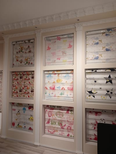 This image captures a close-up, overhead view of a display of rolled-up fabric blinds, likely in a retail setting in the city of Türkmenabat, Turkmenistan. The blinds are organized into three rows, with four blinds visible in the top and middle rows, and five in the bottom row. Each blind features a different cartoon-themed pattern, suggesting they are intended for children's rooms.
The top row displays blinds with patterns of cartoon characters, potentially animals, and sports-related imagery like stars and soccer balls. The middle row showcases blinds with fairytale princesses, butterflies, and other delicate designs. The bottom row continues the theme of cartoon characters, including what appear to be Angry Birds, cacti, and other playful motifs. The blinds are set against a neutral, off-white background, which is part of the store's display fixtures. The overall impression is one of a bright and cheerful children's decor section.
The lighting suggests an indoor environment, possibly a shop during daylight hours, although no specific time of day or weather conditions can be determined. The overhead angle and the way the blinds are presented indicate a merchandising display designed to showcase the variety of patterns available. There is no visible text that can be clearly deciphered, other than the potential word "style" and "icon" on one of the middle-row blinds, which is not sharp enough to be certain. The setting is likely a fabric store or a home decor retailer.