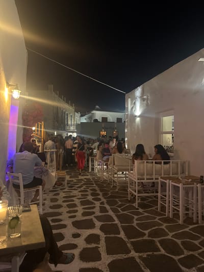 The image depicts a bustling outdoor dining scene at night in Naousa, Greece, likely at a restaurant or bar called "San Fos" as indicated by a sign. White-washed buildings with architectural details line a narrow cobblestone alleyway that serves as a pedestrian thoroughfare and outdoor seating area. The lighting is atmospheric, with warm lights from wall-mounted fixtures and illuminated signage contrasting with the dark night sky.

Numerous patrons are seated at tables and standing around, engaged in conversation and enjoying their evening. Some individuals are dressed in casual attire, while others appear to be dressed for a night out. The seating arrangements include white metal furniture with white cushions, as well as straw-topped stools and tables. In the foreground, a table is set with a drink and a lit candle, suggesting a personal dining experience.

The cobblestone ground is a prominent feature, laid with irregularly shaped stones set in white mortar, creating a distinctive pattern. Red bougainvillea is visible on a trellis, adding a touch of color to the otherwise monochromatic architecture. The overall atmosphere is one of lively social activity and a vibrant nightlife scene.