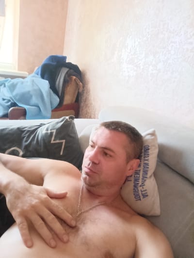 The media file shows a man lying on a sofa, partially undressed. He has short brown hair and is looking to his left with a neutral expression. His right hand is resting on his chest. A small, light-colored pillow with dark blue text is visible behind his head. In the background, there are clothes piled up on what appears to be a window sill or shelf, and a textured wall with a soft peach hue. The scene suggests a relaxed, indoor setting, likely a living room. The lighting indicates it might be daytime, possibly late afternoon, given the soft ambient light. There is no indication of the weather or a specific event. The visible text on the pillow is in Cyrillic script and appears to be an inscription or label.