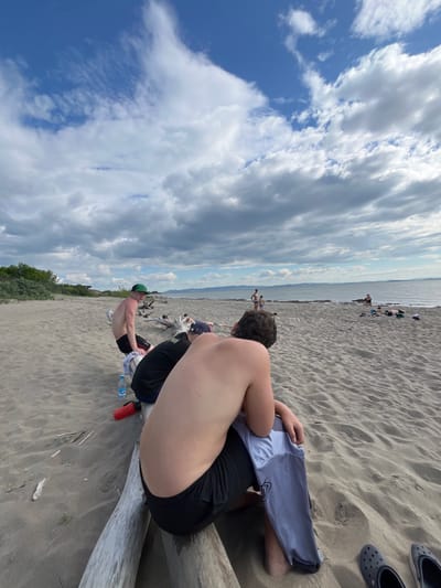 The image depicts a beach scene with several people in the foreground and background. In the foreground, a young man with dark curly hair is sitting with his back to the camera, wearing black shorts and holding a light gray t-shirt. To his left, another young man with fair skin and dark hair is also seated on a log, wearing black shorts and a black t-shirt. He is wearing a green baseball cap. A blue water bottle and a red thermos are visible on the sand near them.
In the mid-ground, a group of people are visible near the water's edge. Some are standing, and others appear to be lying down on the sand. The beach is sandy, with driftwood logs scattered around. The background shows a body of water, possibly a lake or sea, with hills or mountains in the distance under a cloudy sky. The overall environment appears to be a natural, outdoor setting, likely a beach. The specific location in Krasnoturansk, Russia, is not visually identifiable from the image content alone.