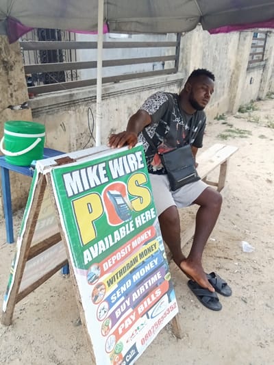 A man wearing black shorts and a gray and black shirt is seated on a wooden bench with his right leg crossed over his left. The bench is situated under a gray and pink umbrella. He has a black shoulder bag slung over his left shoulder. The man is in front of a wooden A-frame advertising stand with a bright green sign that reads, "MIKE WIRE POS AVAILABLE HERE". The sign lists services including "deposit money", "withdraw money", "send money", "buy airtime", "pay bills", and a phone number, "08025658352". The stand is positioned in front of a small wooden table on which a green bucket sits. The environment is an open-air area with a sandy surface and concrete sidewalks and a metal fence in the background. The location is Sapele, Nigeria, at coordinates 5.86 latitude and 5.701 longitude.
