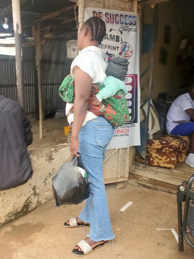 A full-bodied, medium-shot, outdoor, candid photograph captures a woman from the side, facing left, in what appears to be Ohit, Nigeria. She is wearing light blue flared jeans and a white short-sleeved shirt. Her dark hair is styled in braids. A baby, wrapped in a green and orange patterned cloth and a light blue knitted sweater, is being carried on the woman's back. The woman's right hand is holding a black plastic bag. She is wearing decorative, bejeweled sandals on her feet.

Behind her, a large white banner with text in English is visible, including the words "DE SUCCESS Enterprise" and a phone number. The environment suggests an outdoor market or commercial area, with corrugated metal siding and wooden structures in the background. Sunlight illuminates the scene, casting shadows on the sandy ground. A person in a white shirt and blue shorts is partially visible sitting on a patterned cushion to the right of the frame. The overall impression is of a busy, informal urban setting.