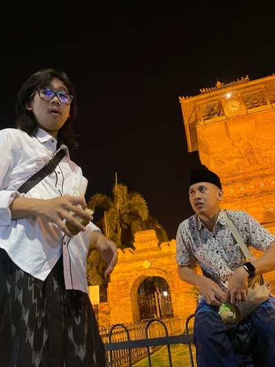 The image depicts two males at night in Kauman, Indonesia, specifically near the Great Mosque of Kauman (Masjid Raya Kauman) complex in Yogyakarta. The background features a large, orange-illuminated traditional Javanese architectural structure, including a prominent clock tower with a visible clock face indicating approximately 10:20-10:25, and an arched gate. Several palm trees are visible between the individuals and the architecture. A dark, low metal fence separates the foreground from the background structure.

The individual on the left, a young male with long dark hair and blue-rimmed glasses, wears a white collared shirt with black trim and a dark sarong with a light geometric pattern. He holds a translucent container with a yellowish liquid at chest height. His mouth is slightly open, and he looks upwards and to the right. A black shoulder bag strap crosses his chest.

The individual on the right, a young male with short hair and a black *peci*, wears a short-sleeved patterned shirt and dark blue patterned pants or sarong. He is seated or crouched on the low metal fence. A light-colored shoulder bag is slung across his chest, and a dark smartwatch is visible on his left wrist. His hands are positioned near his lap, one holding a small green object. He looks upwards and to the left with an alert expression. The overall scene is lit by artificial lighting, predominantly warm orange tones illuminating the architectural background.