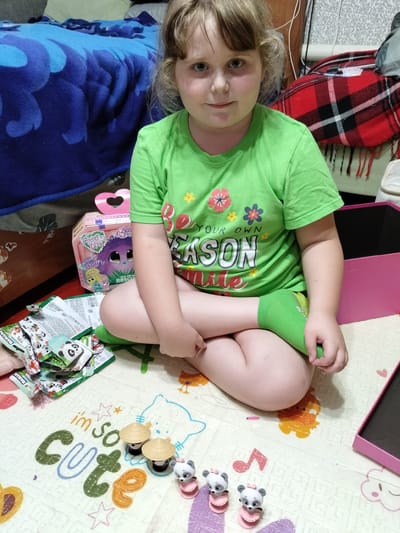 A young girl with blonde hair and blue eyes sits cross-legged on a mat on the floor, facing the camera. She is wearing a bright green t-shirt with floral designs and the text "YOUR OWN KASON Mile" printed on it. She is also wearing green socks.

The mat she is sitting on has a colorful, cartoonish design featuring animals and the words "im so cute." Scattered around her are several small, individually wrapped packets, some of which have "ПАНДЫ" (Pandas) printed on them, suggesting they are collectible toys. There are also three small panda figurines, each wearing a pink dress, and two small straw hats.

Behind the girl, to her left, is a blue and purple blanket with a large graphic on it. To her right is a pink box. The background shows parts of a room, including what appears to be a bed with a red and black plaid blanket draped over it, and patterned wallpaper.

The lighting suggests it is daytime, and the overall scene conveys a sense of playfulness and childhood. The girl's expression is neutral, but the presence of toys and the "cute" inscription on the mat indicate a theme of play. The location context provided is Luhansk, Ukraine.