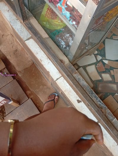 The scene depicts a high-angle view, likely at a storefront or kiosk in Ugbowo, Nigeria. In the foreground, a dark-skinned forearm, adorned with a gold-colored bracelet, extends into the lower right, holding a small, light-colored cylindrical object. A bare foot, with painted toenails, wearing a black flip-flop with a light blue strap, rests on a tiled surface to the left. This tiled surface consists of irregular brown and grey segments. A low, diagonal structure, constructed from a metal frame and a concrete or tiled base, separates the immediate foreground from a background display area. Behind this structure is a glass display case containing various commercial goods. Observable items within the display case include a yellow plastic bag printed with "THANK THANK THANK Have A Good Day," along with multiple packaged items, some in clear plastic. The floor beyond the display case is a mosaic of irregularly shaped, light-colored tiles.
