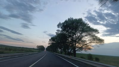 The image captures a view from a moving vehicle on a road near Sokolovo, Bulgaria. A two-lane asphalt road with a solid white centerline stretches into the distance. Metal guardrails line both sides of the road. Rows of trees stand along the right side of the road, blurring due to motion. The landscape on either side of the road features fields. The sky is overcast with various cloud formations, and the light suggests it is either dawn or dusk. A faint light, potentially from an approaching vehicle, is visible further down the road. The overall scene conveys a sense of travel through the countryside.