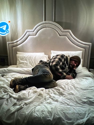 A man lies curled on a bed with a decorative headboard and white bedding. He wears a beanie, a plaid shirt, jeans, and work boots. The room has gray walls. The perspective suggests the viewer is looking down at the scene from above and to the side. A translucent play button with an orange circle is at the bottom left of the image.