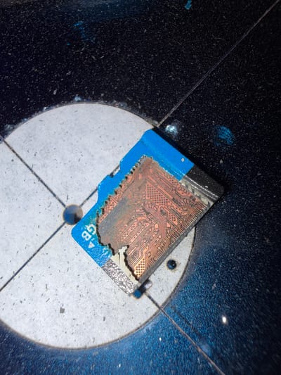 An SD card with a broken blue plastic casing and exposed copper circuitry rests on a light-colored, textured surface. The SD card is positioned diagonally, with its circuitry facing upwards and slightly towards the right. The surface it lies on appears to be circular with two intersecting lines, possibly a base or a stand. Surrounding this surface is a dark, textured area with numerous small, light-colored specks, suggesting dust or debris. The lighting in the image creates reflections and shadows, particularly on the dark surrounding area. The scene is devoid of any people or overt actions. The location is identified as Grapevine, United States of America, though this context does not directly inform the visual description of the object and its immediate surroundings.