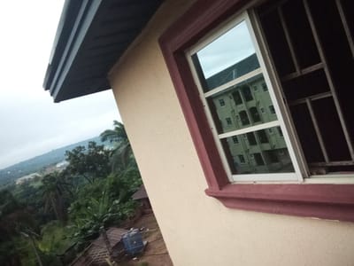 A close-up, low-angle shot captures a portion of a light-colored building in Nanka, Nigeria, with a wide view of the surrounding landscape. The building features a dark grey, corrugated roof overhang on the upper left, extending over a textured, cream-colored wall. A window, framed by a maroon molding, is prominent on the right side. The windowpane reflects a pale green, multi-story building with numerous windows. The reflection suggests the window is positioned on an upper floor of a building.
Beyond the building, the scene transitions to a lush, green environment. Dense tropical vegetation, including banana trees and palm trees, blankets the rolling hills. In the middle ground, a few structures with corrugated metal roofs are visible, along with a blue water tank. In the distance, more buildings are scattered across the landscape, nestled among the hills under a cloudy sky. The overall impression is of a rural or semi-rural setting within the city of Nanka, Nigeria, characterized by verdant hills and modest architecture.