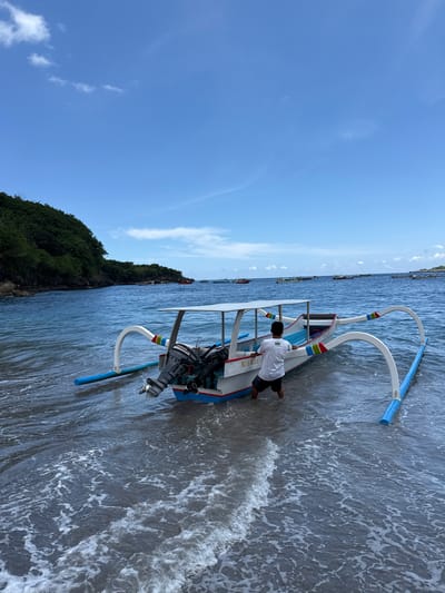 A medium outdoor shot, rotated ninety degrees counterclockwise, depicts a man standing in the shallow ocean next to a beached, small motorized outrigger boat. The man is wearing a white shirt and dark shorts. The outrigger boat is white and blue and has a small Yamaha motor. The bow reads "BLI6N026". The outriggers have blue flotation devices affixed to them. The water is gently lapping at the boat's hull. In the background, there is a tree-covered hillside and other boats are visible offshore. Above is a blue sky with a few puffy white clouds. This scene is located in Sakti, Indonesia.