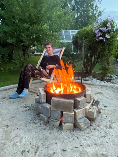 A woman sits in a striped lounge chair next to a fire pit in a yard. She is wearing a black top, black lace pants, blue sandals, and socks. She is holding a bottle of beer. The fire pit is made of metal and surrounded by bricks. There are trees and a greenhouse in the background. The woman appears to be relaxed and enjoying the fire. The overall scene suggests a peaceful evening outdoors.