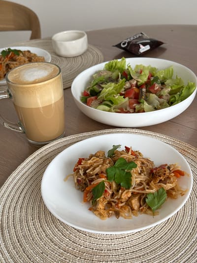 A close-up, eye-level shot showcases a meal and a beverage laid out on a wooden table in what appears to be a domestic setting in Gamovo, Russia. In the foreground, a white plate holds a serving of stir-fried noodles with chicken, carrots, and red peppers, garnished with fresh parsley. To the left, a clear glass mug contains a foamy coffee drink, likely a latte or cappuccino. Behind these, a white bowl overflows with a fresh salad composed of lettuce, tomatoes, and cucumbers, lightly dressed. A dark, individually wrapped snack, possibly chocolate, rests in the background. A small, white ceramic dish sits to the left of the salad bowl. The table is covered with woven placemats under the plate and mug. The overall scene suggests a casual meal enjoyed indoors.