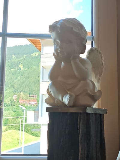 A light-colored cherub statue sits on a dark, rough-textured pedestal in the foreground, positioned to the right of a window. The cherub is depicted in a seated posture, with its head resting on its hands and one wing visible. Through the window, a view of a green, tree-covered mountainside is visible, dotted with buildings and a road. The sky above is bright blue with a few white clouds. The lighting suggests it is daytime. The scene appears to be indoors looking out at a landscape, possibly in the Austrian Alps region based on the provided context mentioning Filzen, Austria.
