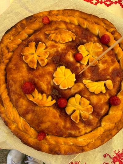 A golden-brown, round pie decorated with braided pastry edges, floral shapes cut from dough, and small, red wild strawberries sits on a beige, textured tablecloth. A skewer is inserted into the pie near the center. The pie appears to be filled with a dark, jam-like substance. In the lower left corner of the image, the tips of white sneakers are visible. The tablecloth has a red embroidered pattern along the right edge. The scene is lit from above, casting shadows on the pie's surface. The location is identified as Вяйнянскі сельскі Савет, Belarus.