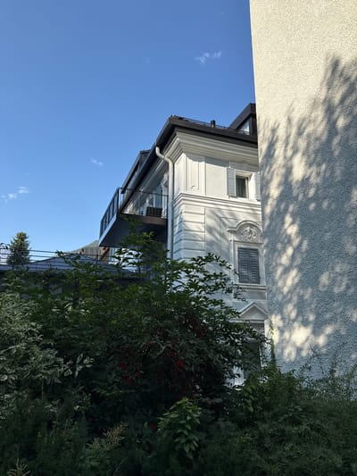 The image shows a portion of a building in Bad Gastein, Austria, with lush green foliage in the foreground. The building is modern with a white facade, black roof, and a balcony with a black railing. It features several windows, some with light blue shutters. The building is partially obscured by the dense greenery, which includes trees and bushes with small red berries. The sky is a clear, bright blue with a few small white clouds. A rough, textured wall of another building casts a shadow pattern from tree branches onto its surface, suggesting the presence of nearby trees. The scene is set during the daytime with bright sunlight.